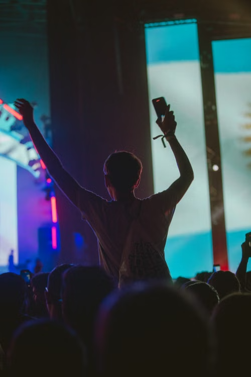 Fan sitting on shoulders with arms raised in front of giant LED stage screens